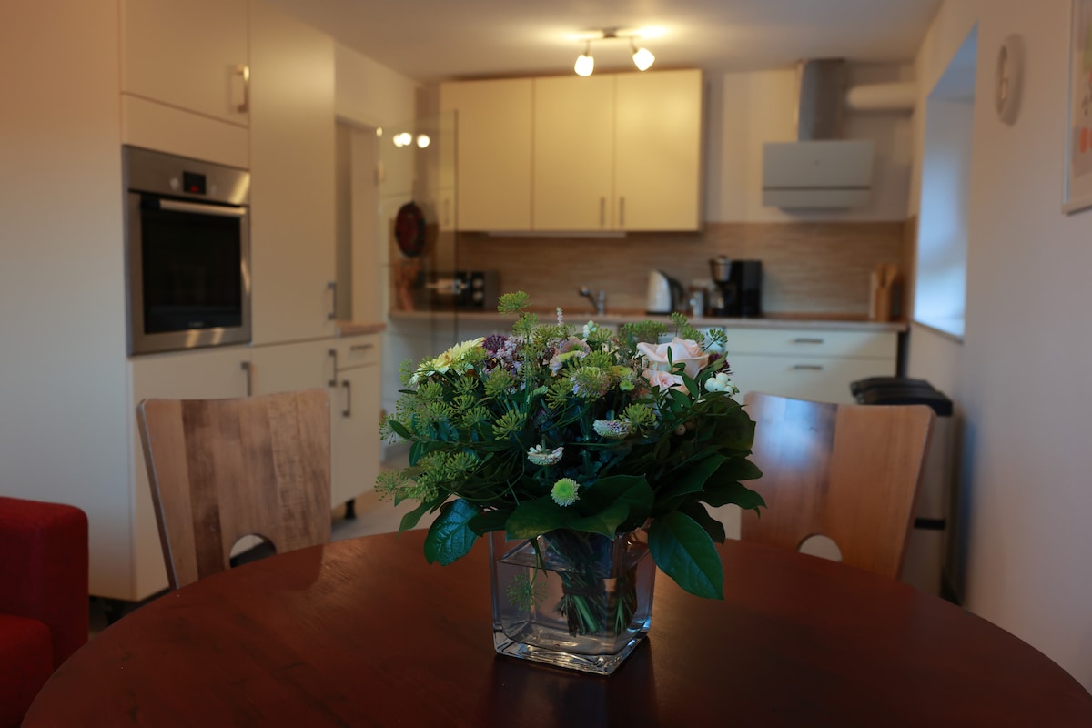 A round wooden dining table is adorned with a vase of fresh flowers, placed center-stage. The kitchen area features modern cabinetry, an oven, and appliances in a neutral color palette, creating a functional and inviting space. Soft lighting enhances the warm atmosphere.