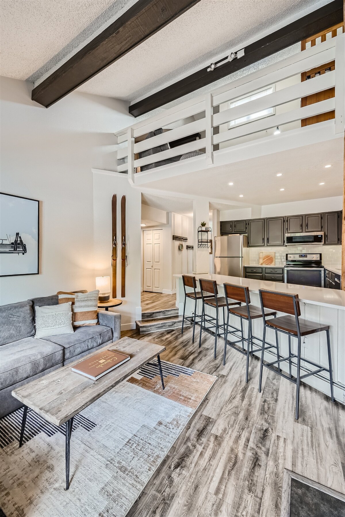 A spacious living area features a comfortable gray sofa, a coffee table, and a patterned area rug. A set of bar stools lines the kitchen counter, which is equipped with modern appliances. Wooden beams add character above, while a pair of skis is displayed on the wall.