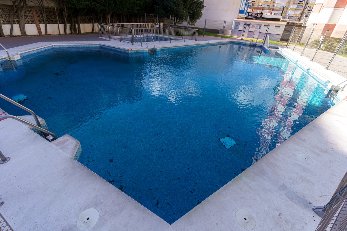 The image displays a large swimming pool with clear blue water, surrounded by a concrete deck and metal railings. The pool's design features steps leading in, creating easy access. Sunlight reflects on the water's surface, enhancing the serene environment of the communal space.