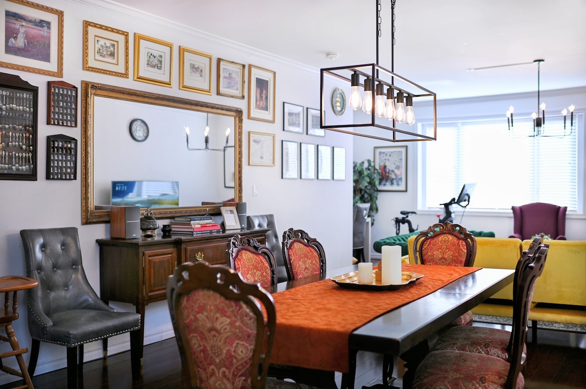 A dining area features a long table set with an orange cloth and a simple centerpiece. Surrounding the table are various chairs, including ornate ones with red upholstery. A large mirror and a collection of framed artworks adorn the walls, while a glimpse of the living area with colorful seating is visible.