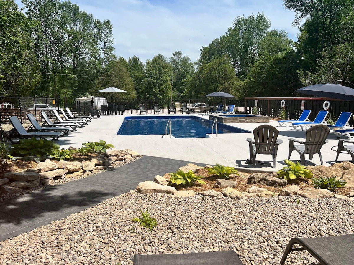 A spacious swimming pool is surrounded by a smooth deck with multiple lounge chairs. Umbrellas provide shade, and a rock garden with greenery enhances the outdoor setting. Trees create a serene backdrop, contributing to a relaxing atmosphere.