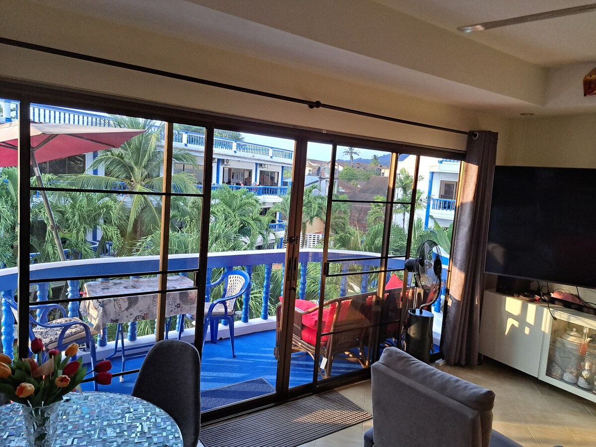 A spacious living area features large glass doors opening to a balcony with views of lush greenery. Comfortable seating is present, and a large TV is mounted on the wall. Light enters through the transparent doors, enhancing the airy atmosphere.