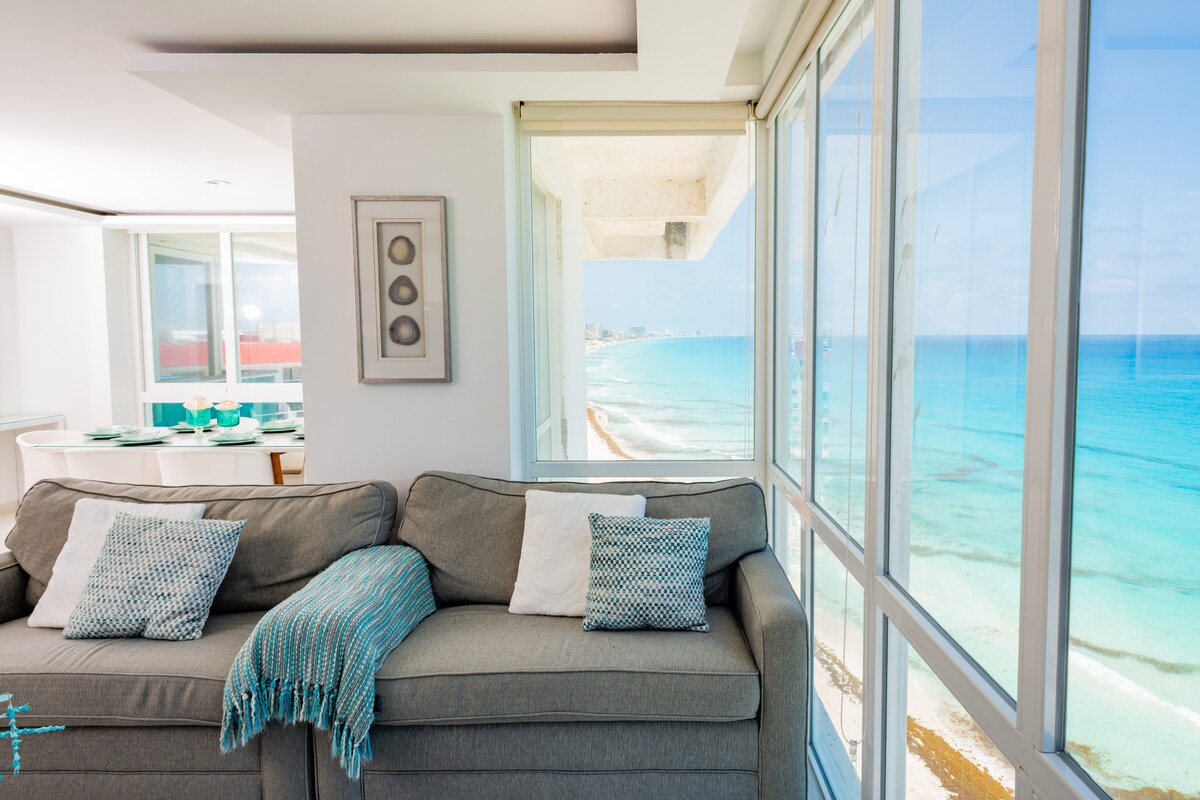 A comfortable sofa positioned to capture stunning views of the turquoise ocean through large glass windows. Natural light enhances the soft neutral tones of the decor, while a light blue throw and decorative pillows add subtle color to the cozy seating area.
