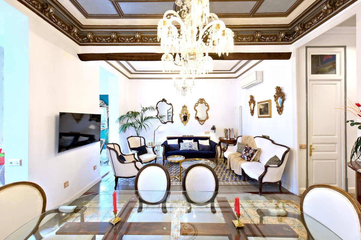 The living area features elegant furnishings, including a dark blue sofa and a light-colored settee, arranged around a patterned area rug. A decorative chandelier hangs from the ceiling, while mirrors are positioned on the walls, enhancing the bright ambiance created by the large window.