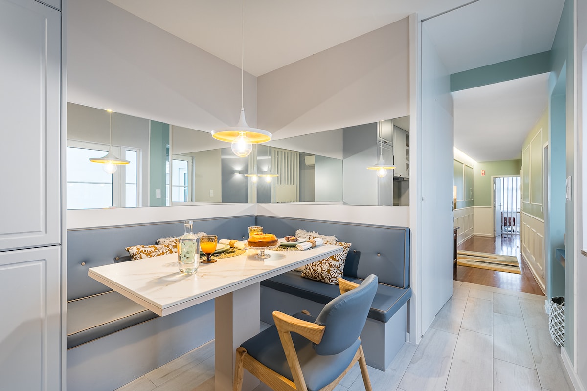 A light-filled dining area features a minimalist table set with refreshments. Comfortable seating is arranged around the table, and a pendant light hangs above, enhancing the clean and modern design. Mirrors along the wall reflect the inviting atmosphere.