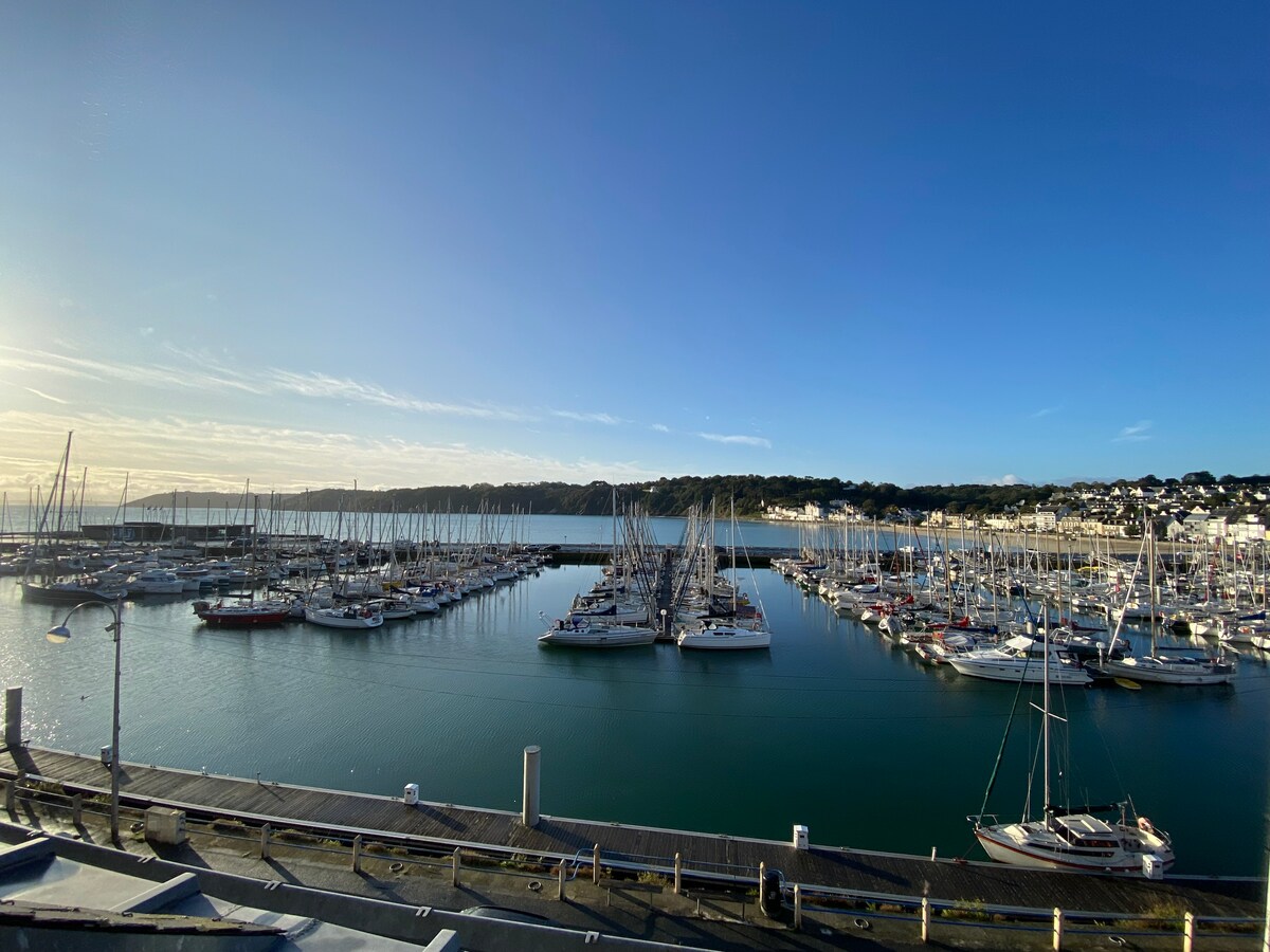 A view of the marina is presented, showcasing numerous sailboats moored in calm waters. Beyond the boats, the landscape reveals gentle hills and a clear blue sky, enhancing the serene atmosphere of the port. The reflection of the boats can be seen on the water's surface.
