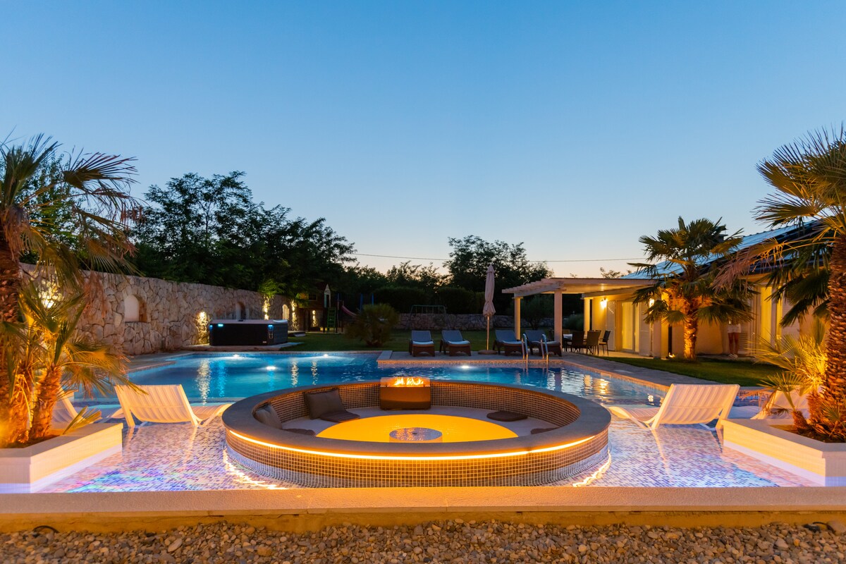 A soothing evening scene features a spacious swimming pool, surrounded by palm trees, with a sunken seating area illuminated by soft lighting. Lounge chairs are positioned nearby, and a covered terrace is visible in the background.