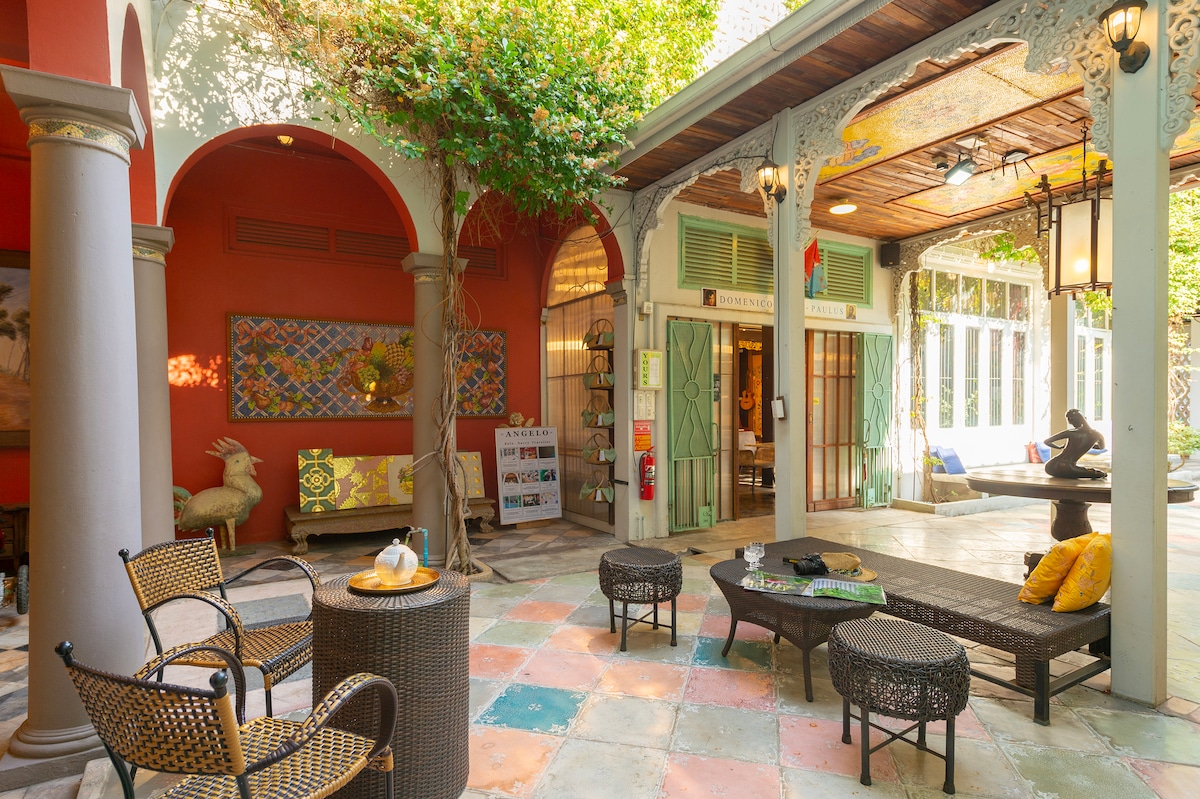 A lively courtyard is framed by arched entries and greenery, showcasing a blend of colorful floor tiles. Seating options include woven chairs and tables arranged invitingly. Artwork is displayed on the walls, enhancing the unique design of the shared space.
