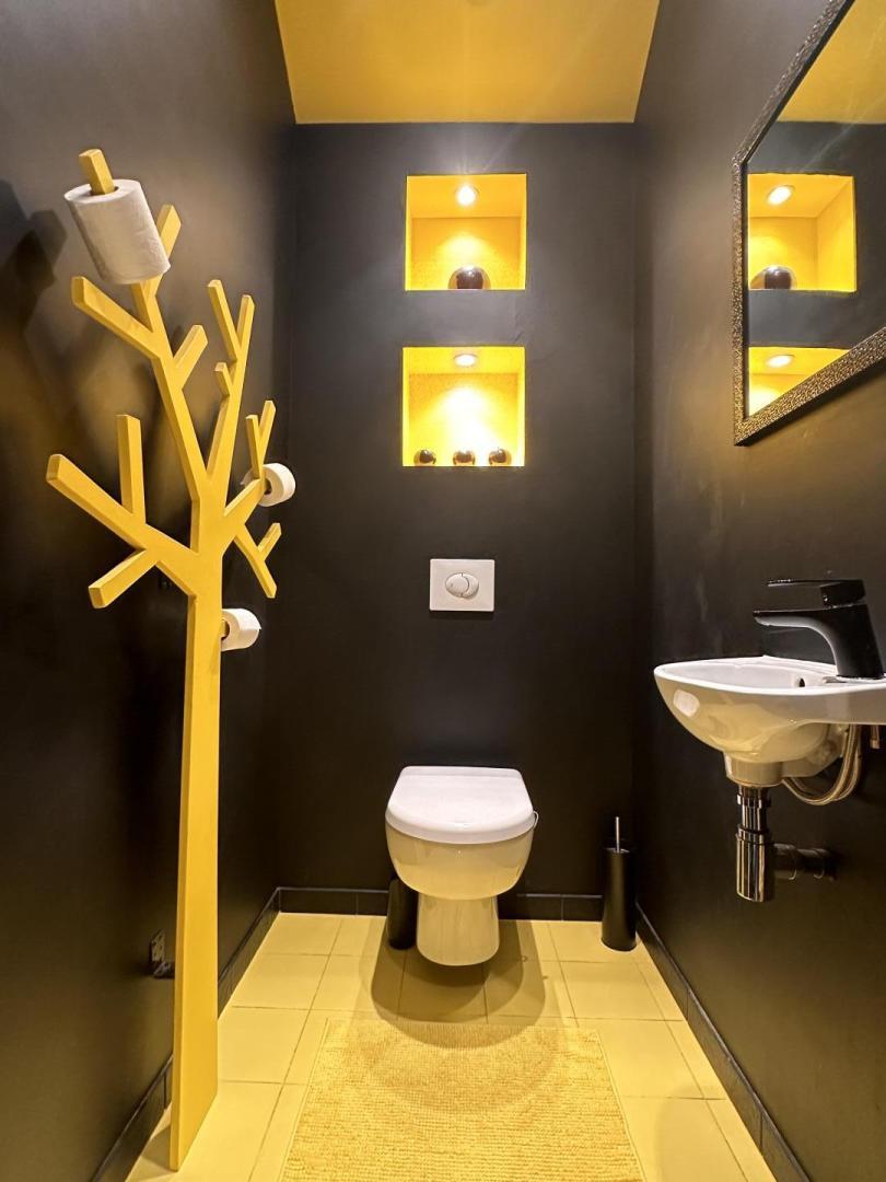 A modern bathroom features a yellow accent wall with illuminated shelves displaying decorative items. A white toilet is positioned beside a sleek black sink. A novel tree-shaped towel holder is mounted on the wall, enhancing the design with functional elegance.