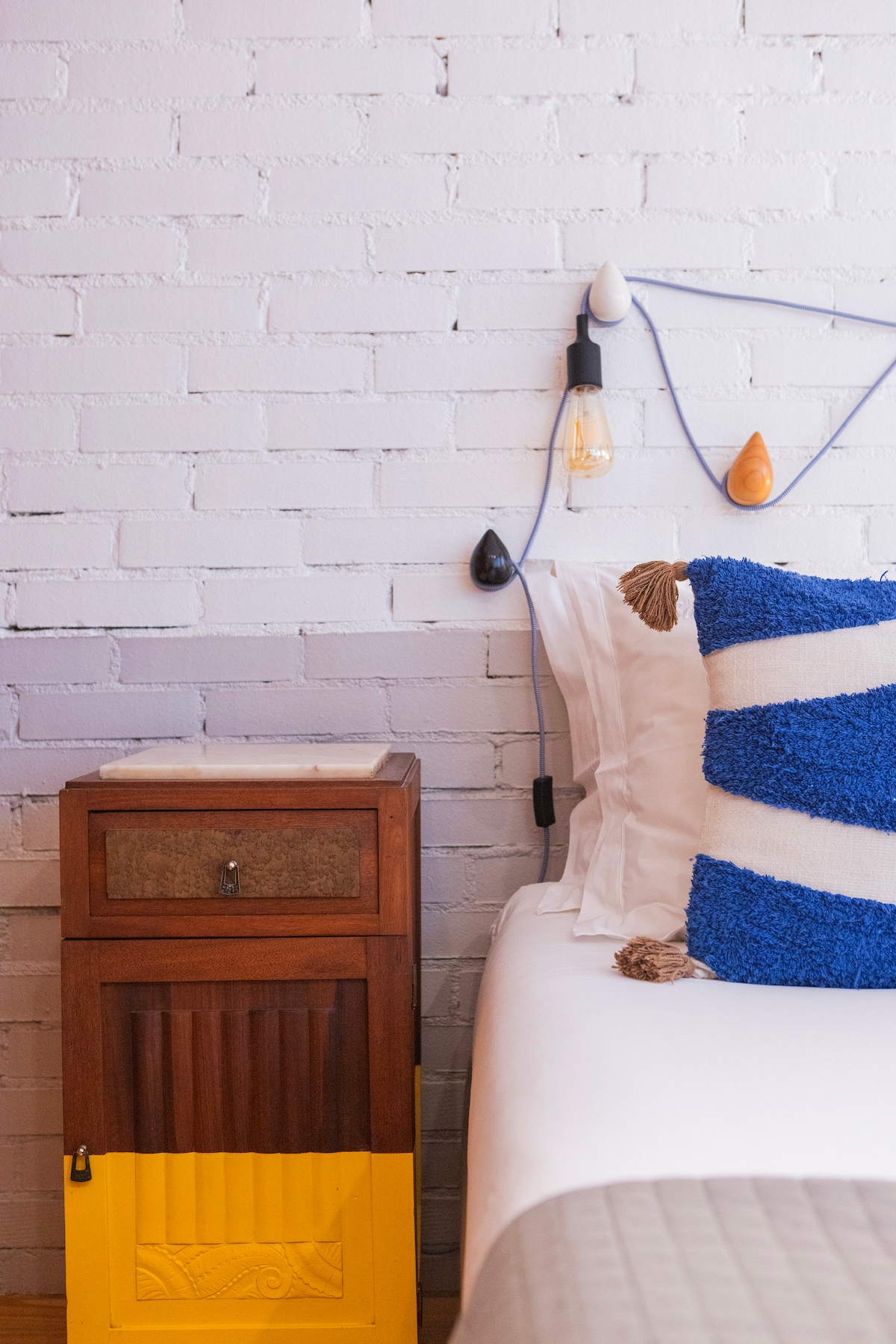 A cozy bedroom corner features a spacious bed with white linens and a textured blue and white decorative pillow. A bedside table, painted in yellow and wood, is complemented by an elegant lamp suspended above, adding a subtle warmth to the space.