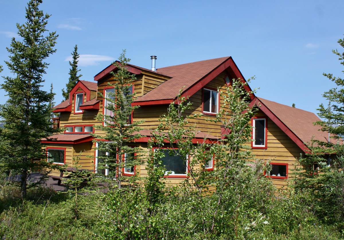The craftsman-built home features a multi-level structure adorned with a combination of red accents and natural wood. Surrounded by verdant foliage and trees, large windows provide glimpses of the landscape. A chimney rises from the roof, indicating a cozy atmosphere within.