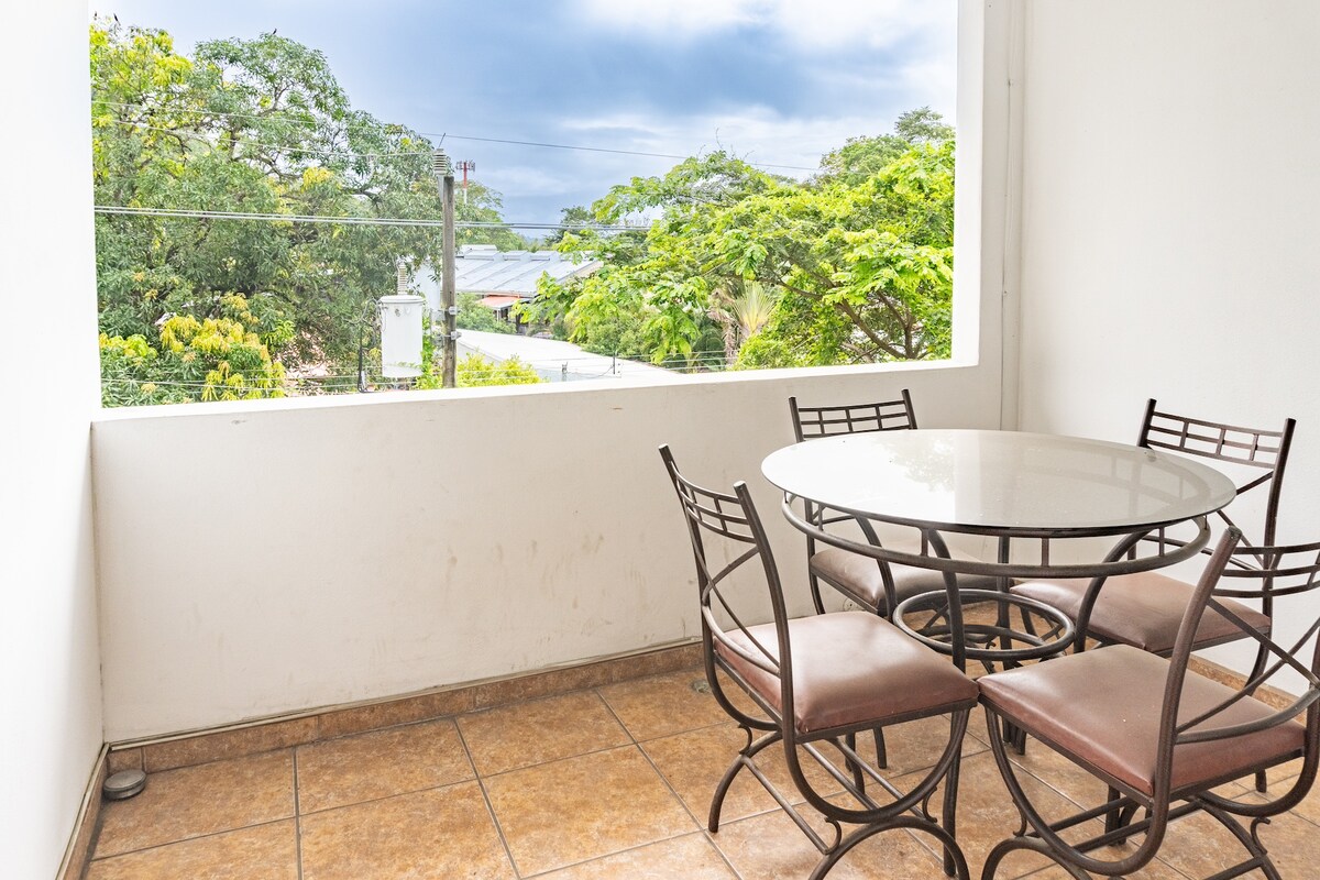 A bright outdoor patio area features a circular glass-top table surrounded by four metal chairs. Lush greenery is visible through the open window, with glimpses of the nearby ocean. The tiled floor enhances the space's inviting atmosphere.