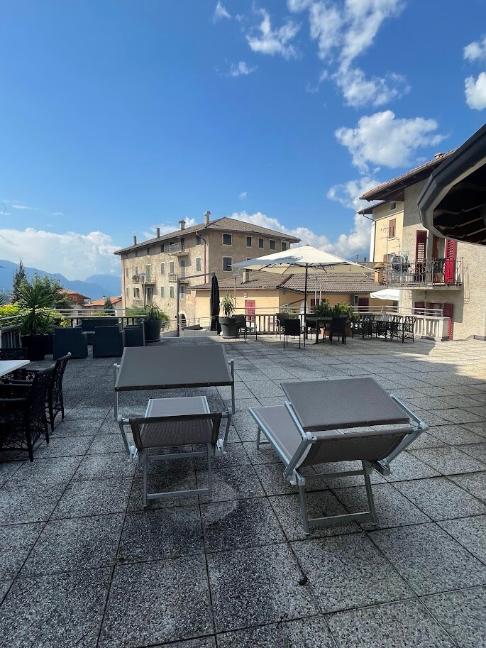 A spacious terrace is depicted, featuring several lounge chairs arranged for relaxation. Umbrellas offer shade over dining tables. Surrounding structures are visible in the background, under a partly cloudy sky that allows sunlight to stream down.