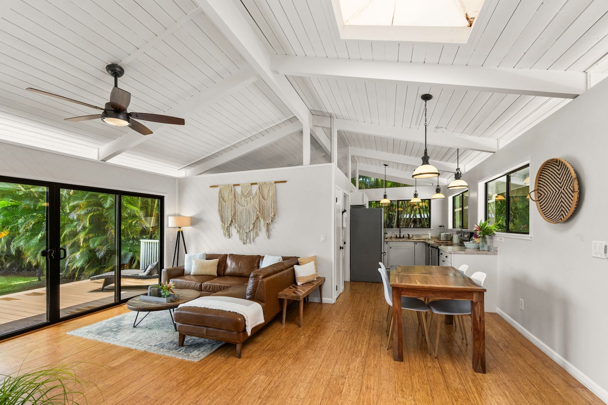 An open concept living space is displayed, featuring a comfortable leather sofa and a wooden dining table. Large sliding glass doors invite natural light, while a modern kitchen is visible in the background. Ceiling fans and decorative elements enhance the airy ambiance.