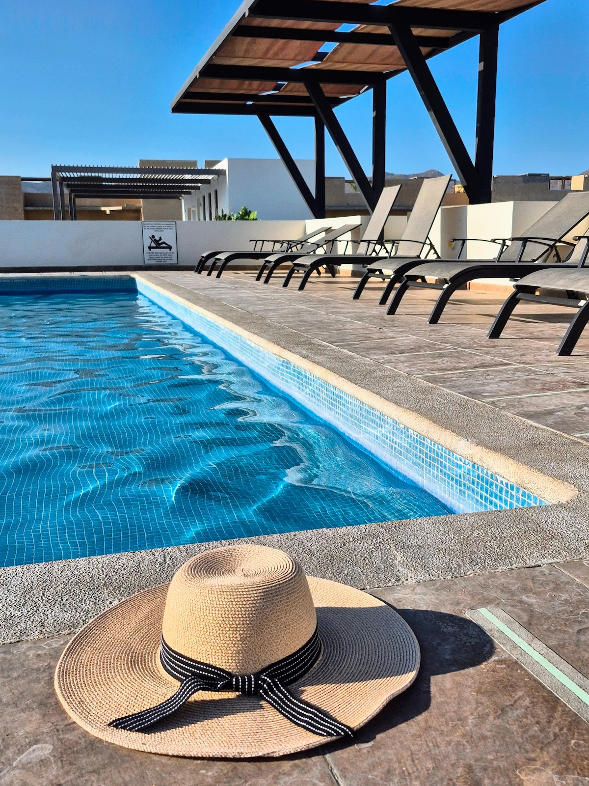A refreshing swimming pool is framed by a sunbathing area with multiple lounge chairs. A wide-brimmed hat rests on the pool deck beside the water, capturing the sunny ambiance of the outdoor space.