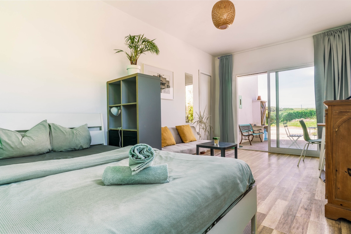 A bright and airy private studio features a spacious bed with gray linens, complemented by neatly rolled towels. A modern bookshelf stands against the wall, while light streams in through large glass doors that open to an outdoor area with seating.