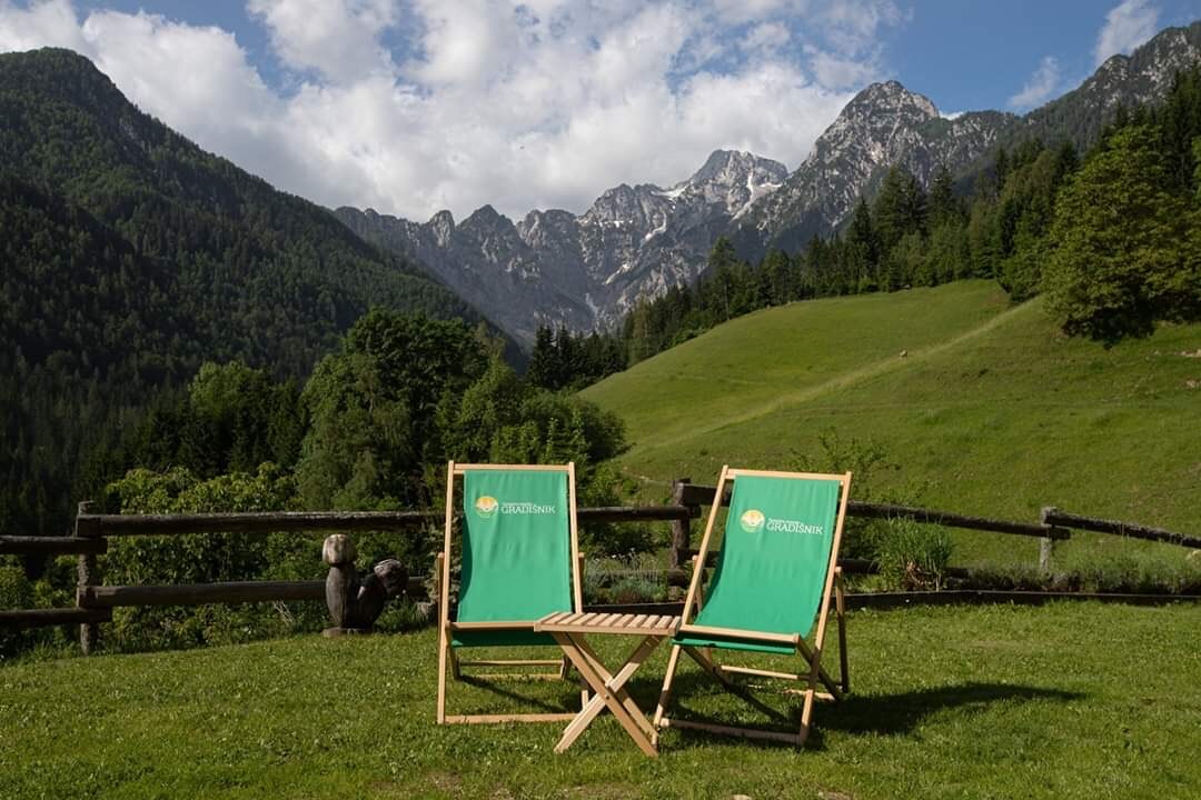 Two green lounge chairs are positioned on a grassy area, facing a stunning view of the mountains. The backdrop features lush green hills and a partially cloudy sky, enhancing the serene landscape surrounded by nature.