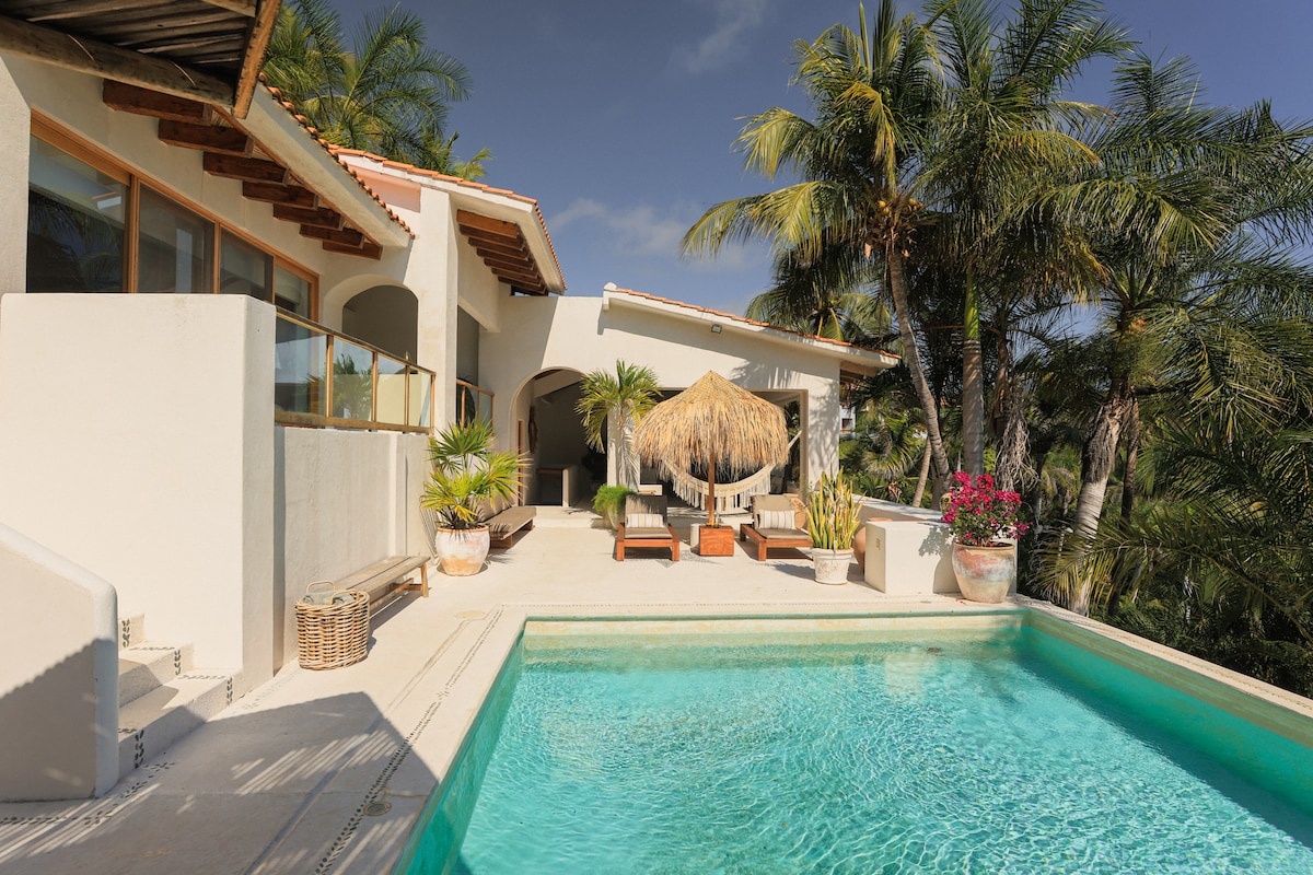 A spacious pool area features a large, clear swimming pool surrounded by palm trees and colorful potted plants. Comfortable loungers and a thatched palapa provide shaded seating, while the villa's warm architecture complements the inviting outdoor space.