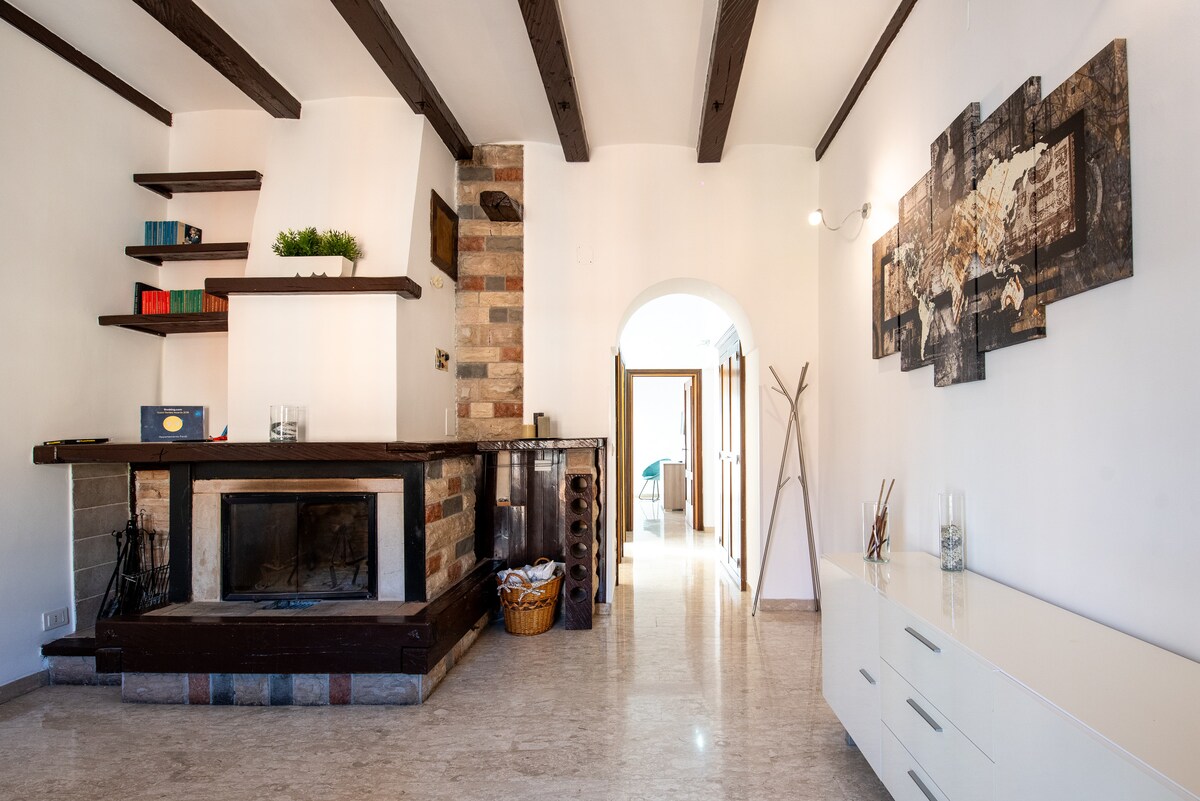 A spacious living area features exposed wooden beams and a stone fireplace, providing a rustic charm. Open shelves display decorative items, while a light-colored sideboard adds storage. A cozy seating arrangement invites relaxation, and a glimpse of the hallway leads to additional rooms.