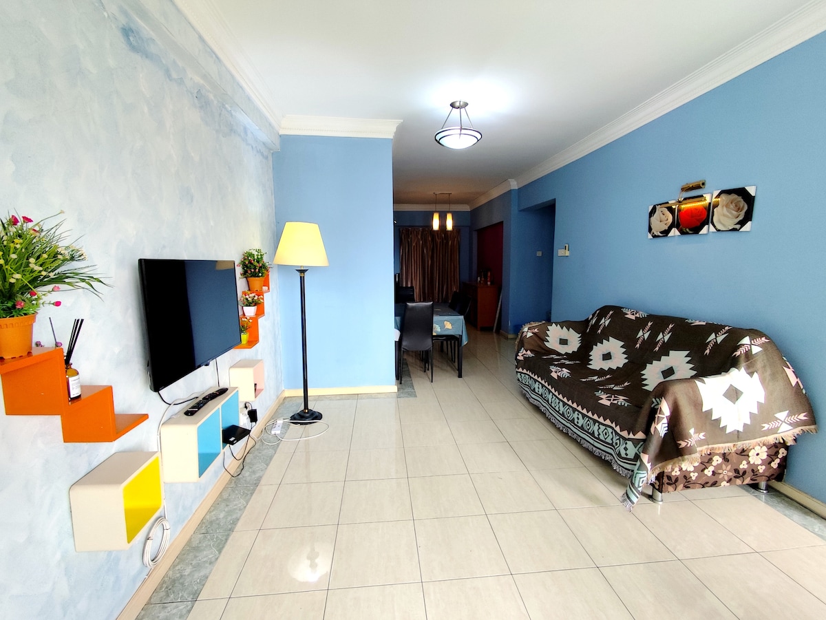 A well-lit living area features a modern design with blue walls and tiled flooring. A comfortable sofa draped in a patterned blanket adds a welcoming touch. A flat-screen TV is mounted on the wall, complemented by colorful shelves displaying decorative items. Dining space is visible in the background.
