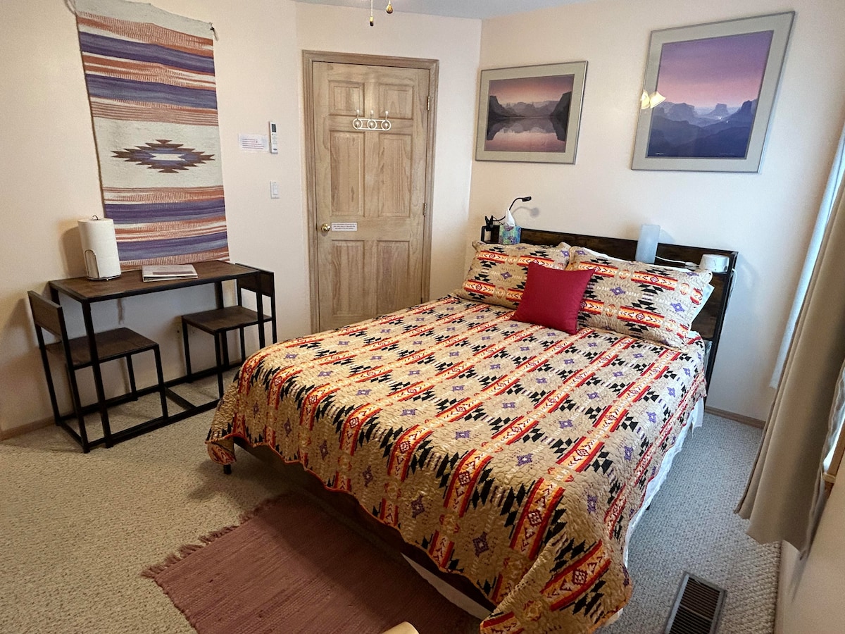 A cozy room features a queen bed dressed in a colorful patterned quilt. A small table with two chairs is positioned beside a door. Wall art complements the southwest decor, while natural light enters through a window covered with light curtains.