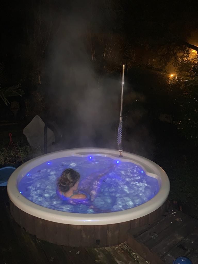A private outdoor jacuzzi is illuminated with soft blue lights, surrounded by a wooden deck. Steam rises from the water, creating a serene atmosphere amid lush greenery in the background. Dim lighting from nearby structures enhances the tranquil setting.