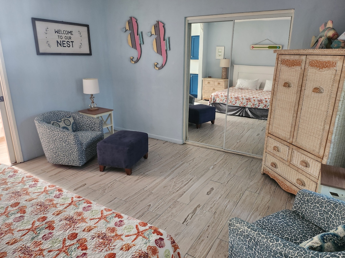 A studio space features a colorful embroidered bedspread and a mirrored closet. Complementary seating includes a patterned armchair and a small ottoman. Soft blue walls and decorative wall art add a cheerful touch, while a bedside lamp provides gentle lighting.
