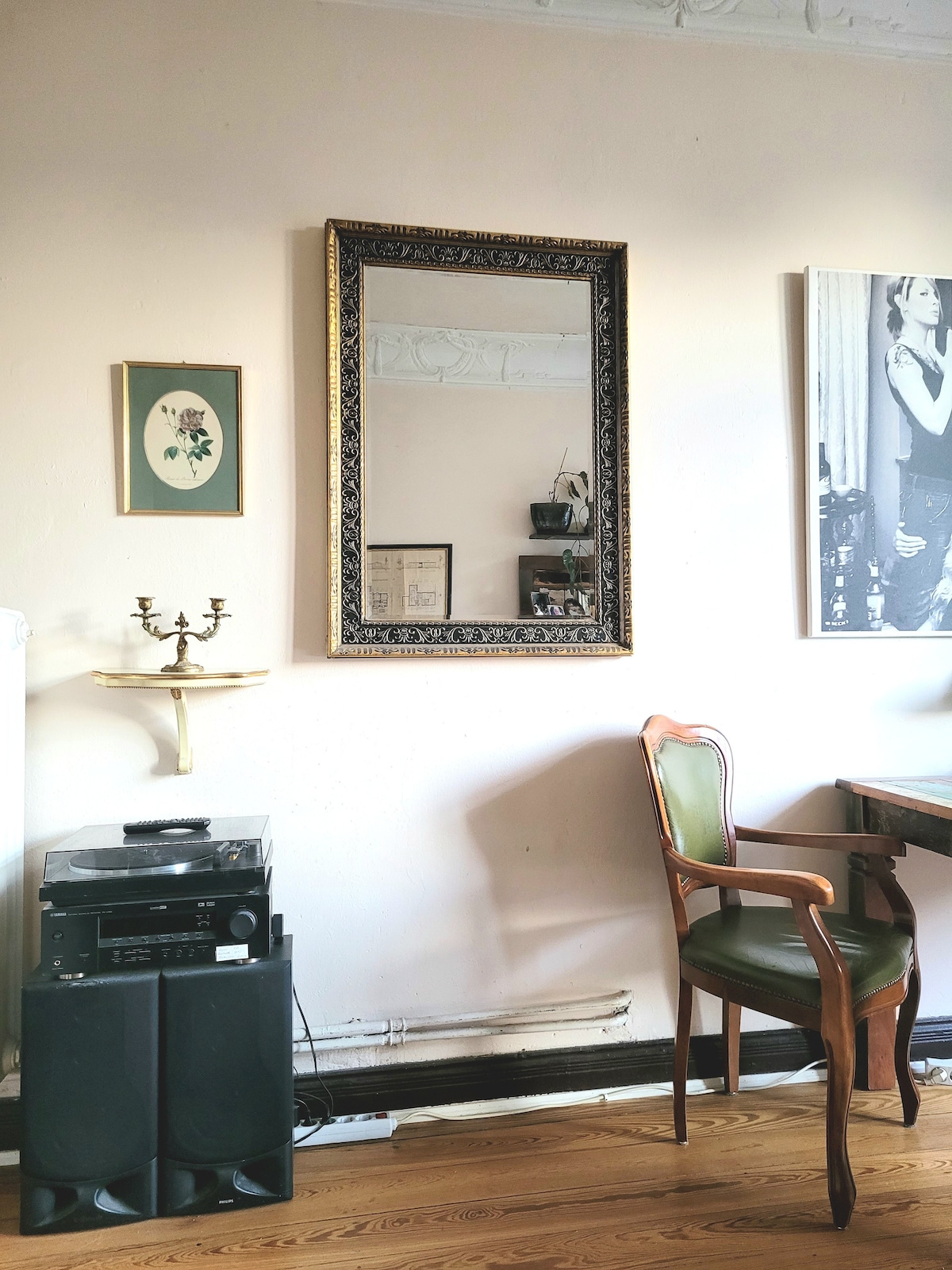 A vintage-style mirror is framed and mounted on a light-colored wall, reflecting the surrounding decor. A wooden chair with green upholstery sits next to a black speaker system. An elegant wall-mounted shelf displays small decorative items, contributing to the room's cozy ambiance.