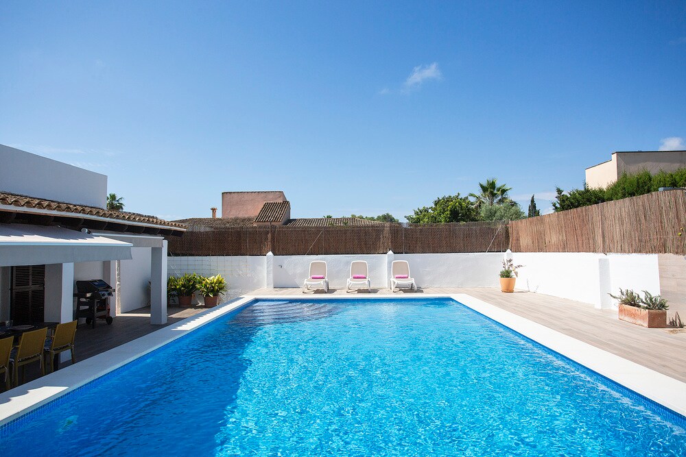 A large swimming pool is featured in the backyard, surrounded by a sun terrace. Three lounge chairs are positioned beside the pool for relaxation. A covered outdoor dining area is visible, reflecting a bright blue sky above and a tastefully landscaped backdrop.