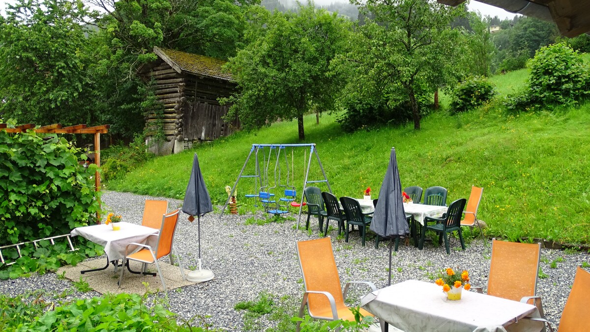 An outdoor seating area features multiple tables with orange chairs and umbrellas, set on a gravel surface. Nearby, a swing set is visible among green grass and trees, with a rustic wooden structure in the background, enhancing the natural surroundings.