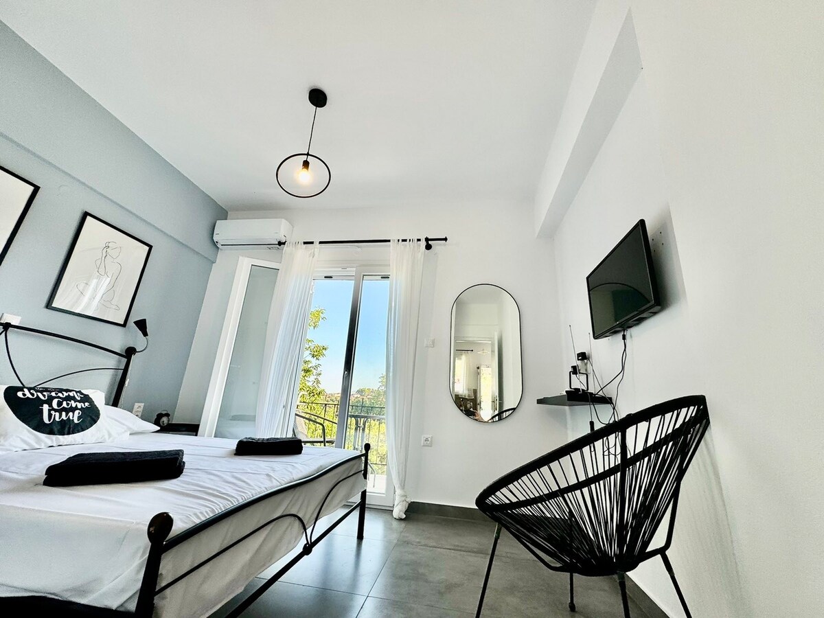 A bright studio features a spacious bed with black linens, complemented by a chic black chair. Natural light filters in through large sliding doors leading to a balcony, and a round mirror adds depth. A television is mounted on the wall, enhancing functionality.