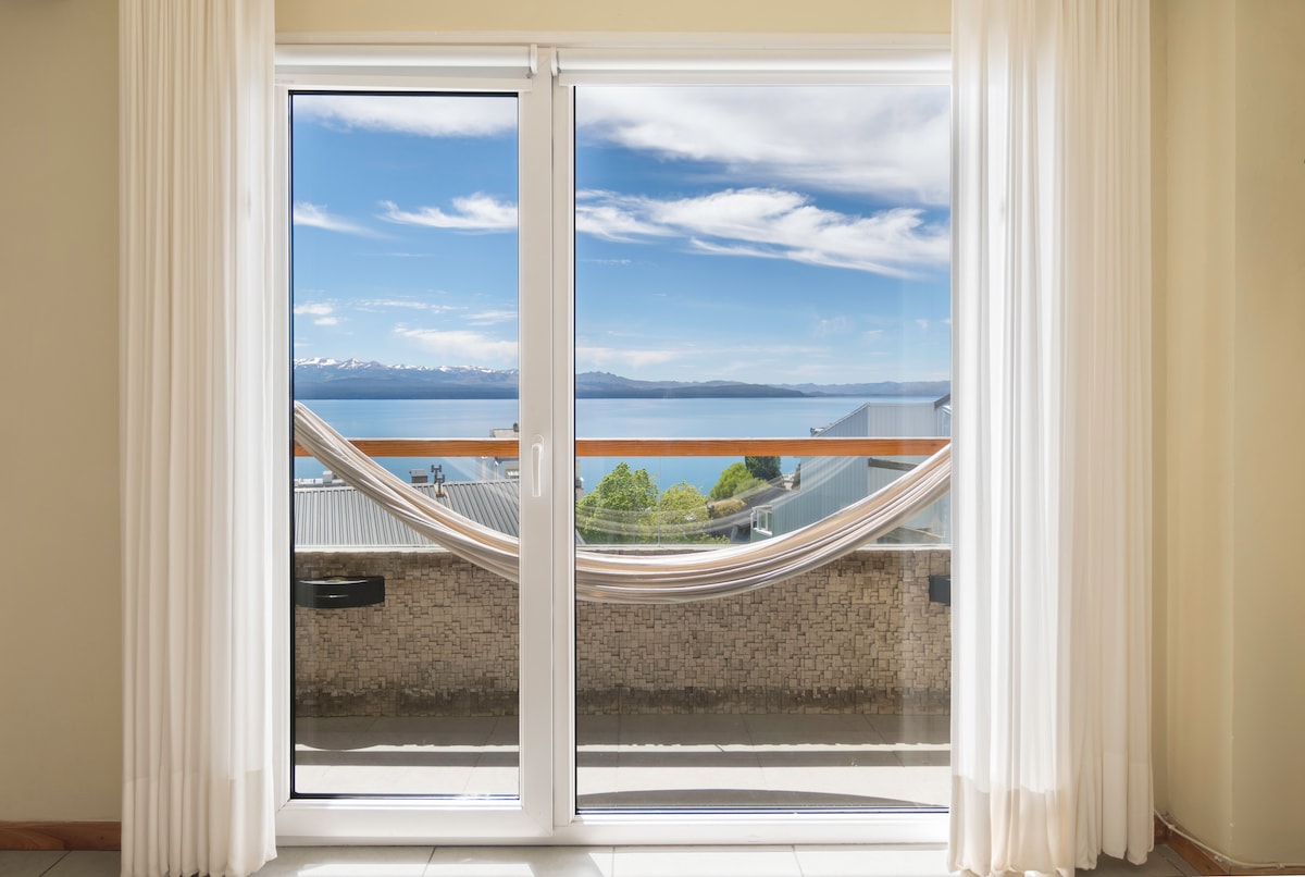 Large sliding glass doors open to reveal a stunning view of Lake Nahuel Huapi, framed by sheer white curtains. A comfortable hammock is positioned on the balcony, inviting relaxation while enjoying the scenery.