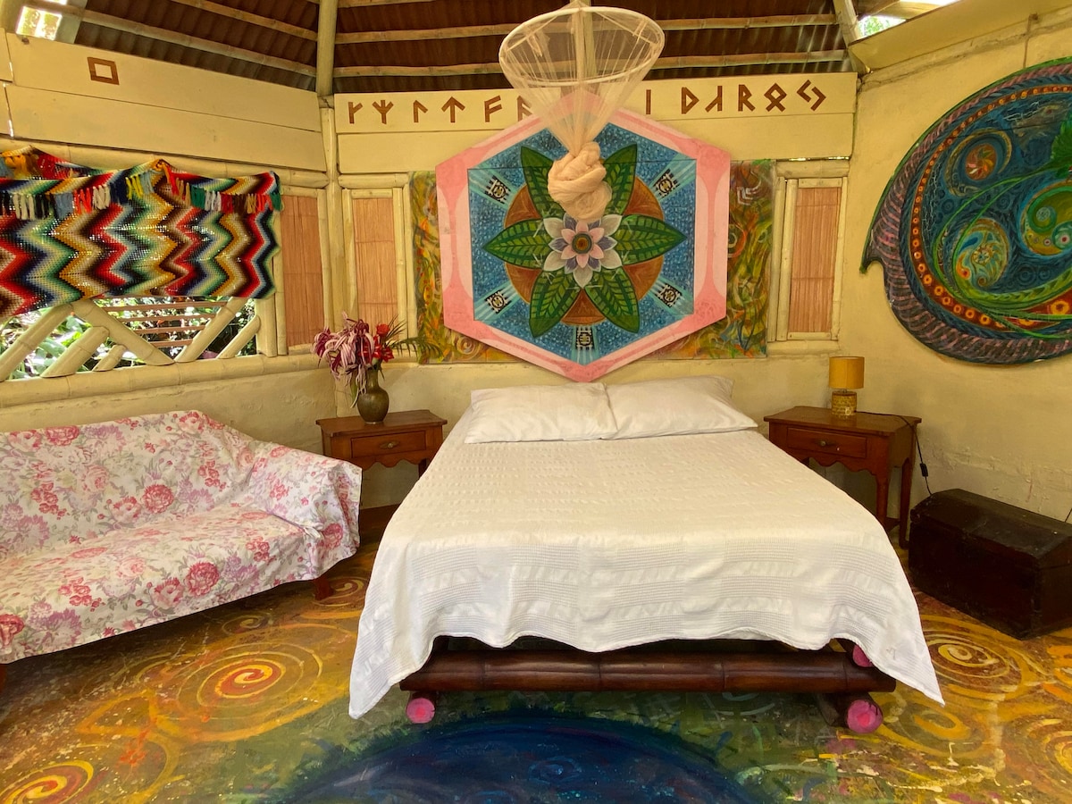 The interior features a hexagonal layout with a spacious bed centered beneath a colorful canopy. A decorative mural featuring a geometric design is displayed above the bed. A floral sofa complements the room, and vibrant patterns adorn the walls, creating an artistic ambiance.