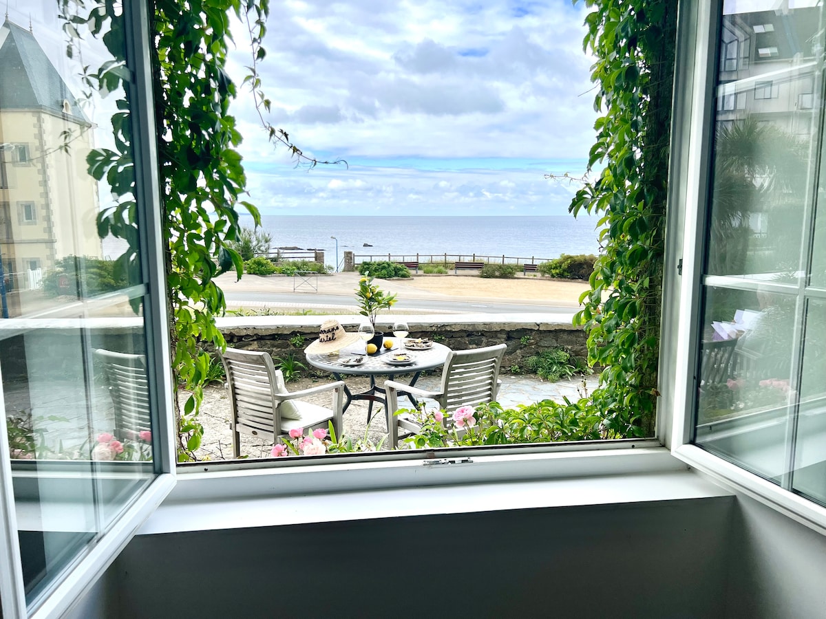 A view from an open window frames a small outdoor table set for two, facing the sea. Lush greenery and flowering plants surround the scene, while the coastline and distant horizon create a serene backdrop under a partly cloudy sky.