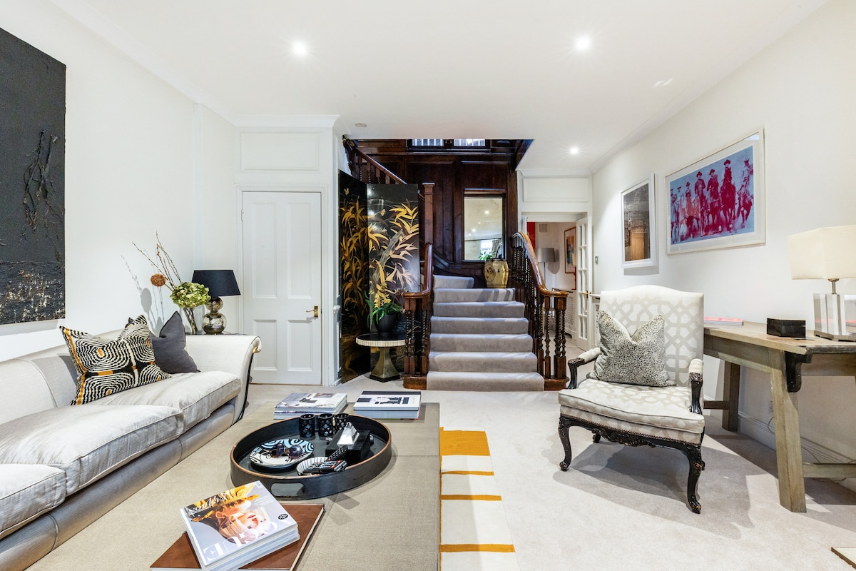 A spacious living room features a comfortable sofa adorned with decorative cushions. A circular coffee table is centered in the room, holding various magazines. Artwork is displayed on the walls, and a staircase leads to the upper floors, enhancing the inviting atmosphere.