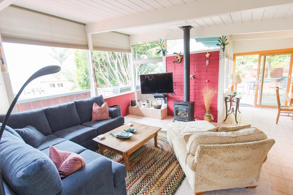 The living area features a large, comfortable sectional sofa and a cozy armchair. A wooden coffee table sits in the center, accompanied by decorative items. A television is positioned on a stand, with a wood-burning stove nearby. Large windows allow natural light to fill the space.