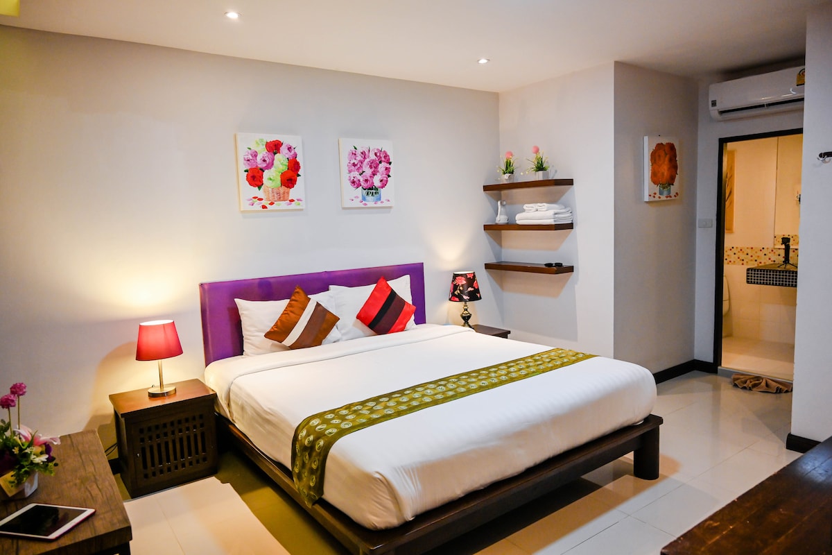 A spacious bedroom is equipped with a large bed adorned with decorative pillows. A bedside table with a lamp complements the room. Shelves display neatly arranged towels and decorative items. A private bathroom is accessible through an open doorway, and air conditioning controls are visible on the wall.