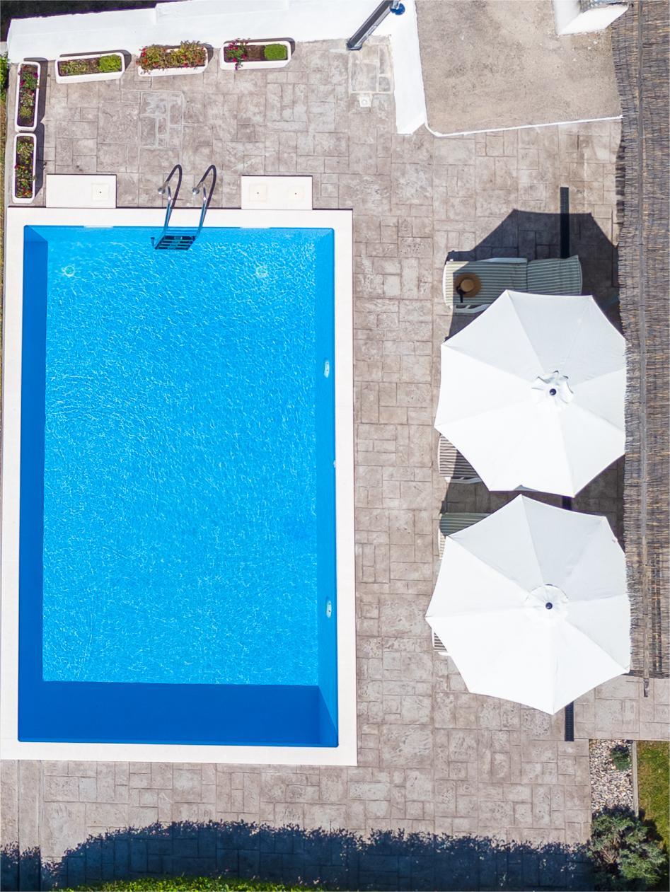 A private rectangular pool with clear blue water is surrounded by a spacious patio. Two large white umbrellas provide shade over sun loungers positioned nearby. The pool area is bordered by neatly arranged flower beds.