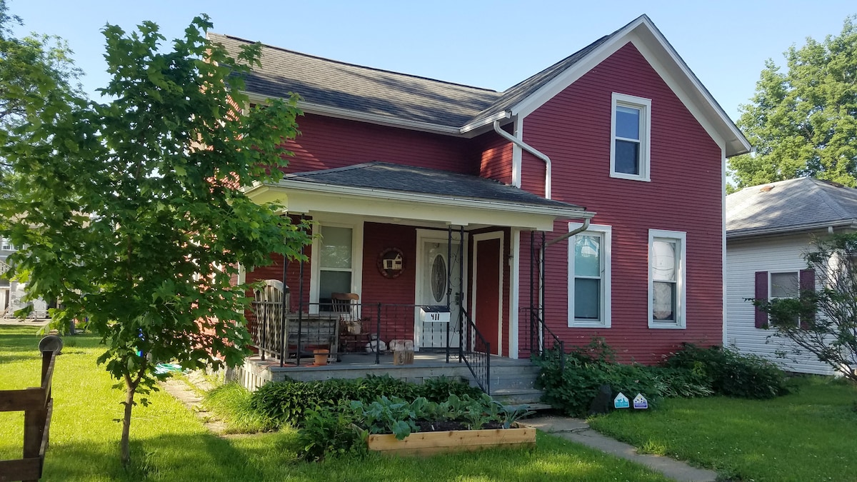 A renovated red farmhouse features a covered porch with seating and a welcoming front door. Lush green grass surrounds the property, complemented by garden beds. Trees provide shade, while the house showcases multiple windows that allow for natural light.