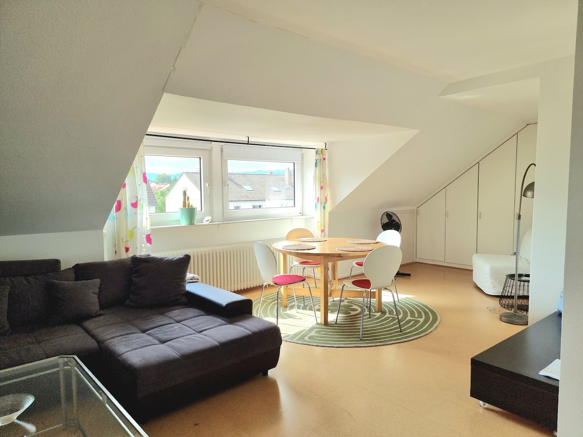 A naturally lit living area features a dark sectional sofa and a round dining table surrounded by white chairs. Large windows are adorned with light, colorful curtains. A rug lies under the table, complementing the overall spaciousness of the room.