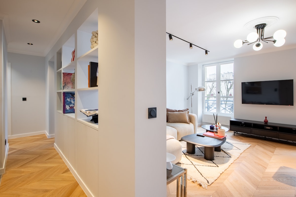 A spacious living area features a light-colored sofa paired with a minimalist coffee table and a soft area rug. Shelving units line the walls, displaying books and decorative items. A television is mounted opposite the seating area, illuminated by natural light from the window.