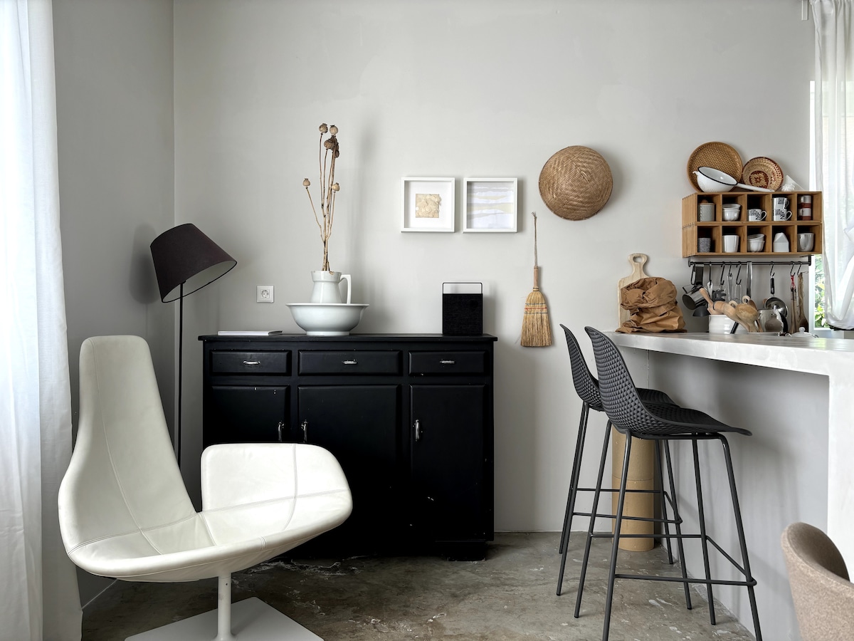 A stylish open kitchen and dining area features a sleek black cabinet and a modern white chair. Decorative items, including woven baskets and framed artworks, are displayed on the walls. A small bar with stools complements the minimalist aesthetic of the space.