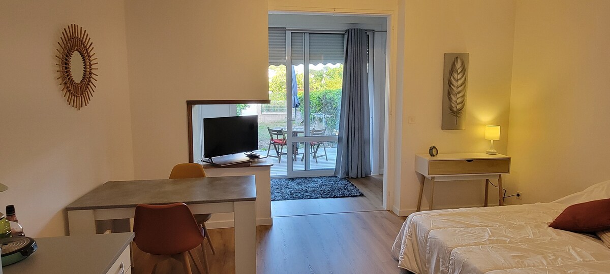 A studio interior is seen, featuring a spacious bed with white linens and a decorative pillow. A desk and chair are positioned near a wall-mounted television. Glass doors lead to a terrace area with outdoor seating, filling the space with natural light.