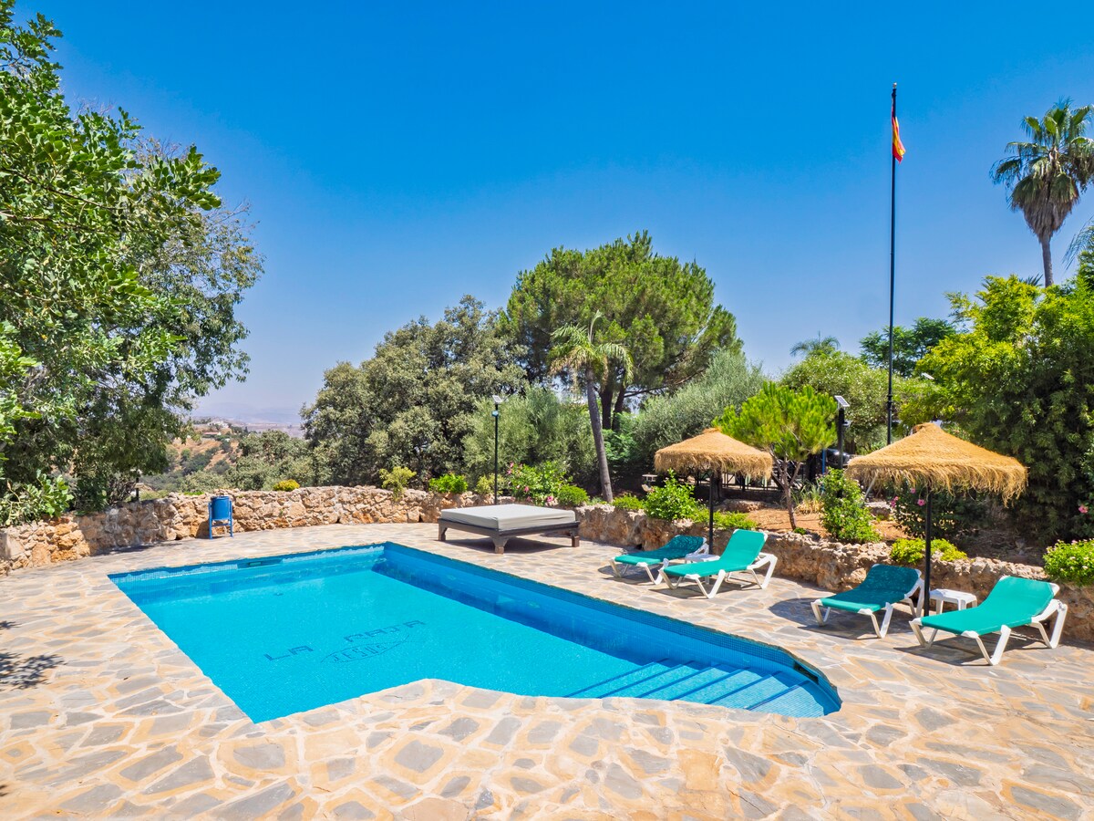 The image displays a private swimming pool surrounded by stone flooring. Lounge chairs with cushions are arranged nearby, and shaded areas are provided by thatched umbrellas. Lush greenery and trees frame the space, enhancing the tranquil outdoor setting.