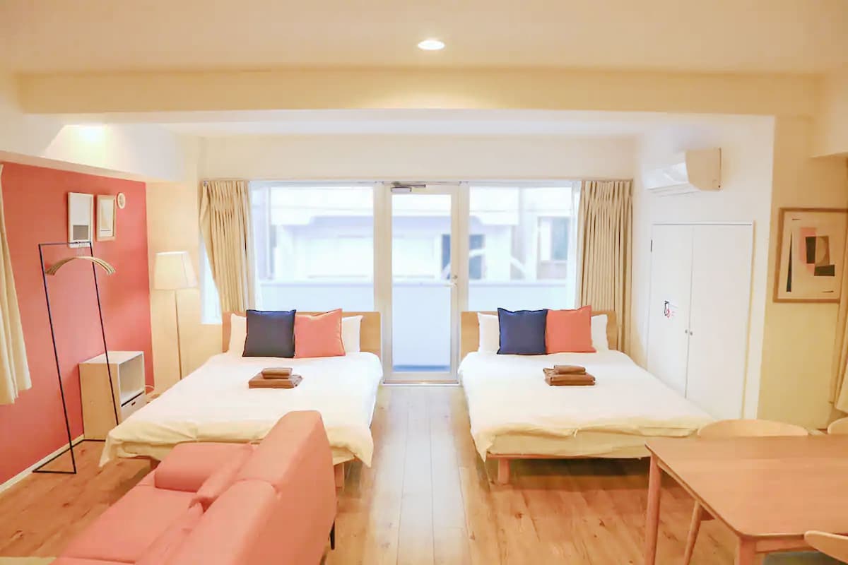 The studio apartment features two spacious beds adorned with colorful decorative pillows. Natural light filters through large windows, illuminating the warm wooden floor. A small dining table and a cozy seating area are also visible, complemented by soft furnishings against the stylish wall accents.