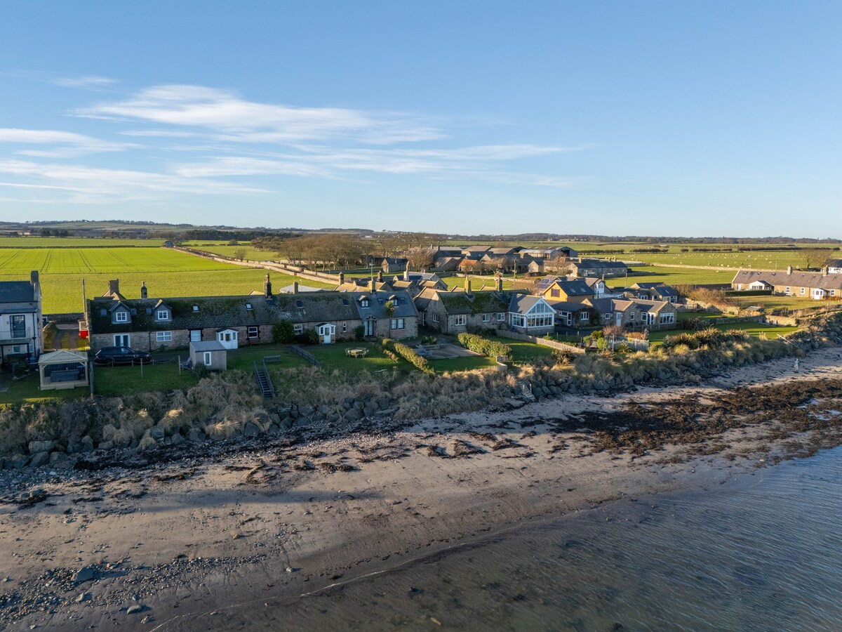 A coastal view captures a row of charming cottages lining the beach, surrounded by serene countryside. The shoreline features sandy areas intermixed with rocks, while fields extend inland under a clear blue sky.