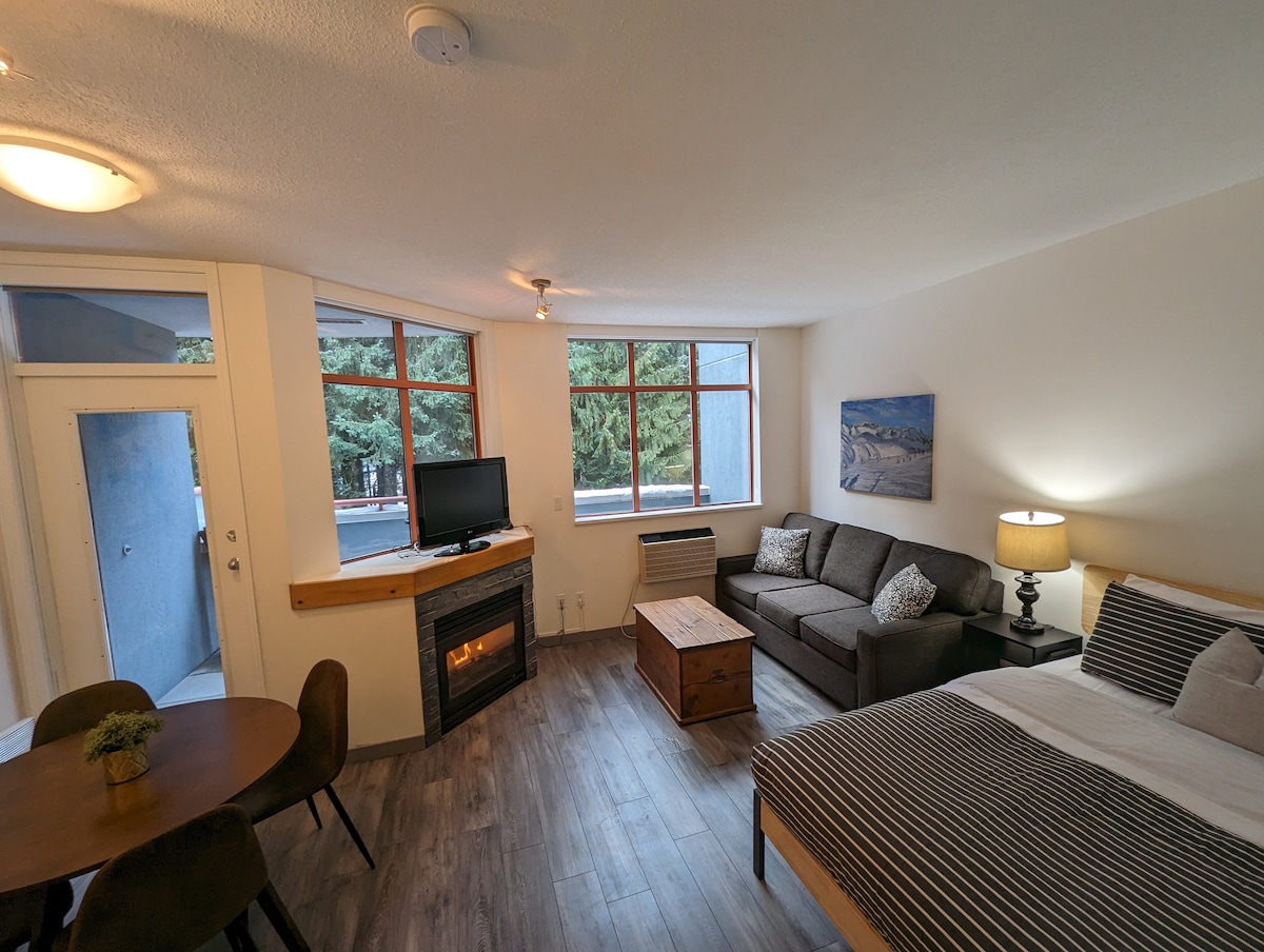 A spacious studio layout contains a cozy gas fireplace and a comfortable seating area with a sectional sofa. Large windows offer natural light and views of nearby greenery. A wooden dining table accommodates four guests, and a TV is positioned for entertainment.