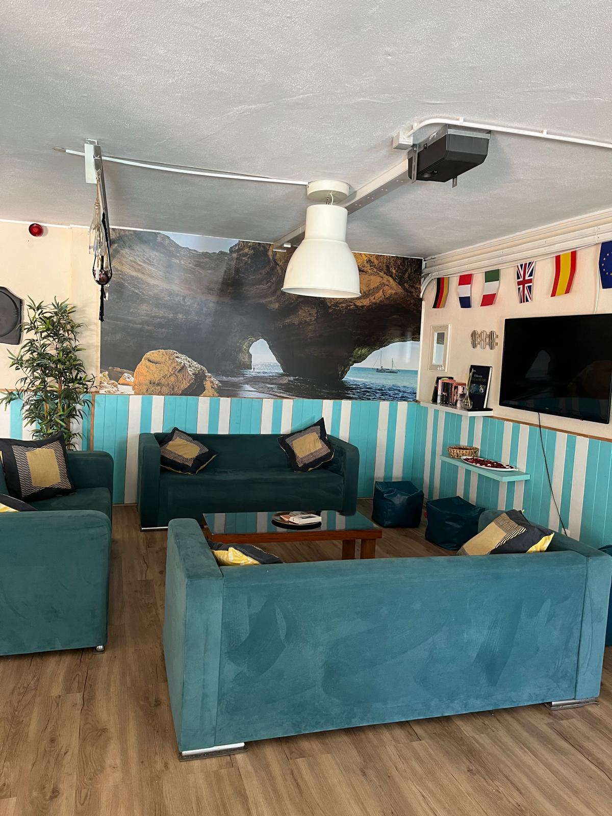 A modern lounge area is furnished with turquoise sofas, accent pillows, and a coffee table. A large coastal mural is displayed on one wall, while a flat-screen TV and decorative items are arranged on another. Natural light is introduced through the ceiling fixture.
