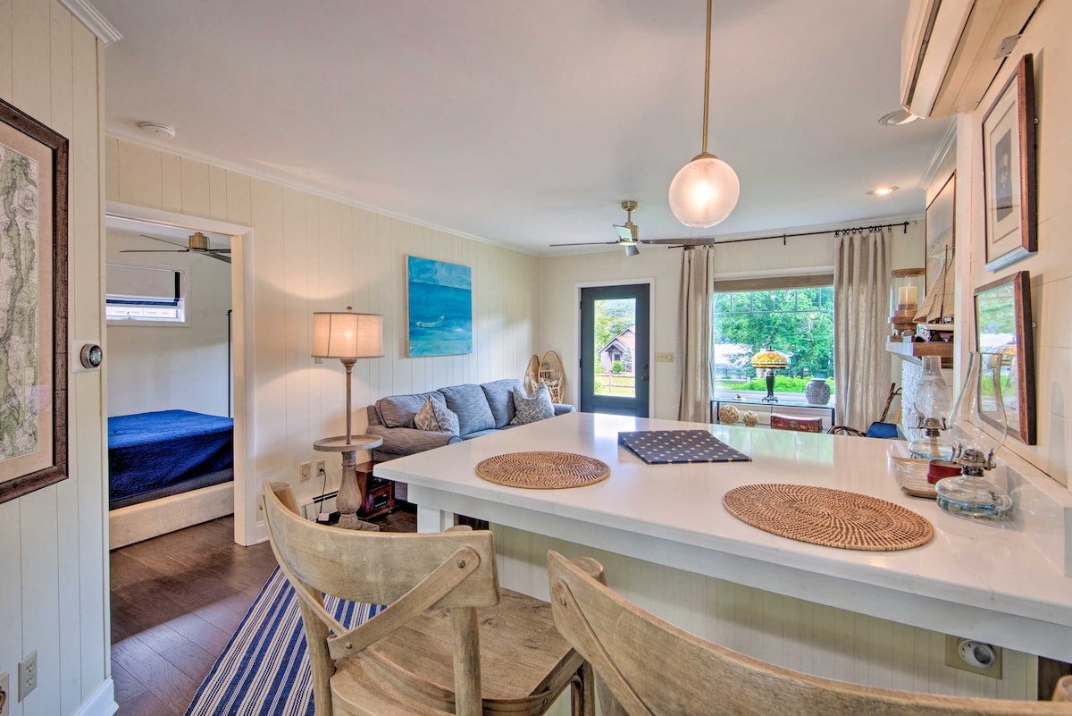 An inviting open-concept space features a well-equipped kitchen with a white countertop and wooden chairs. A cozy seating area is seen with a sofa and nearby artwork. Natural light filters through the windows, illuminating the room, while a glimpse of a bedroom can be seen in the background.