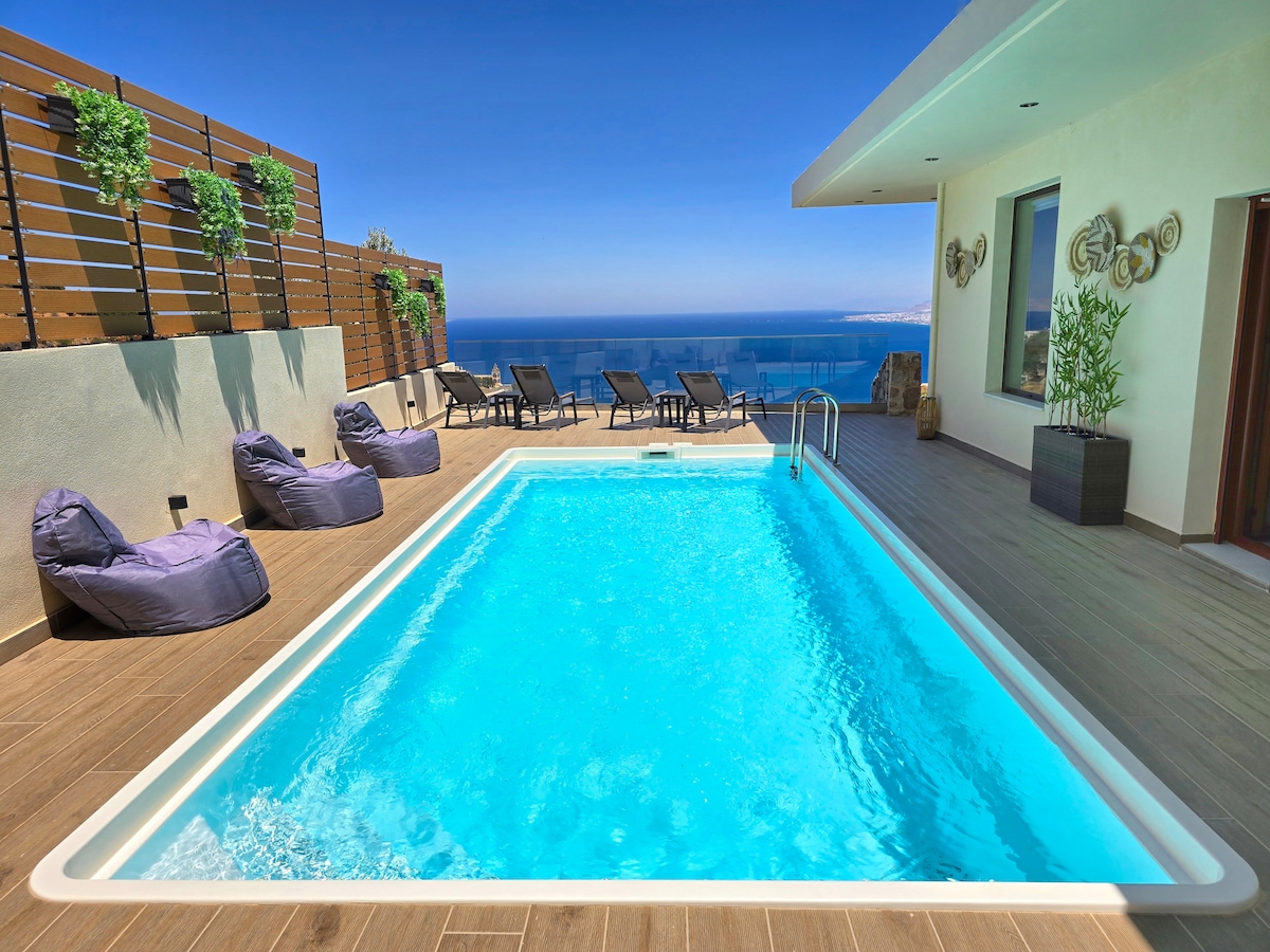 A private swimming pool is surrounded by a wooden deck, featuring several lounge chairs and bean bags for relaxation. The scenic view of the sea and sky enhances the tranquil atmosphere, while the villa's exterior showcases modern architectural elements.